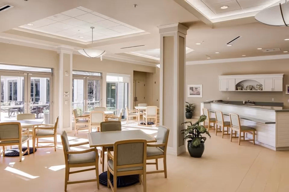 A bright and spacious dining area in a senior living facility with multiple tables and chairs arranged for seating. Large windows and glass doors allow natural light to fill the room. There is a counter with bar stools on the right side, and potted plants add a touch of greenery. The walls are painted in a neutral color, and ceiling lights provide additional illumination.