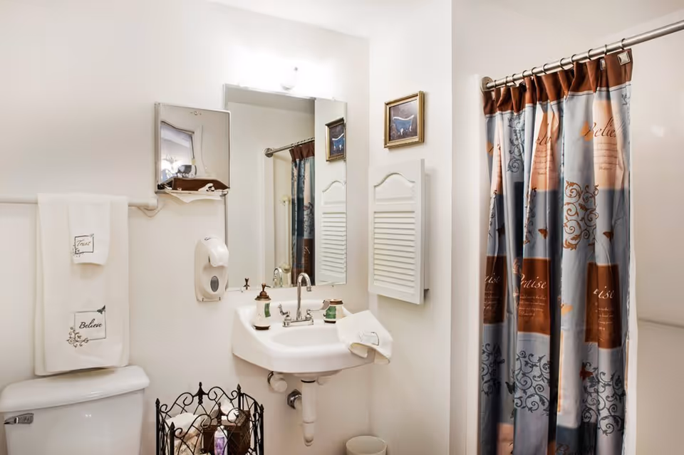 A clean and well-lit bathroom featuring a white sink with a mirror above it, a soap dispenser mounted on the wall, a toilet with neatly folded towels hanging above it, and a shower area with a decorative curtain. There is also a small framed picture on the wall and a metal basket holding toiletries.
