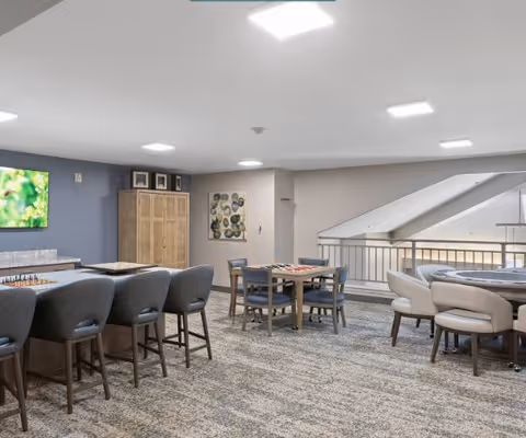 A spacious common area with multiple seating arrangements including a bar-height counter with stools, a square table with chairs, and a round table with cushioned chairs. The room has carpeted flooring, a large TV mounted on the wall, a wooden cabinet, and modern ceiling lights. The area is well-lit and has a neutral color palette with blue and beige tones.