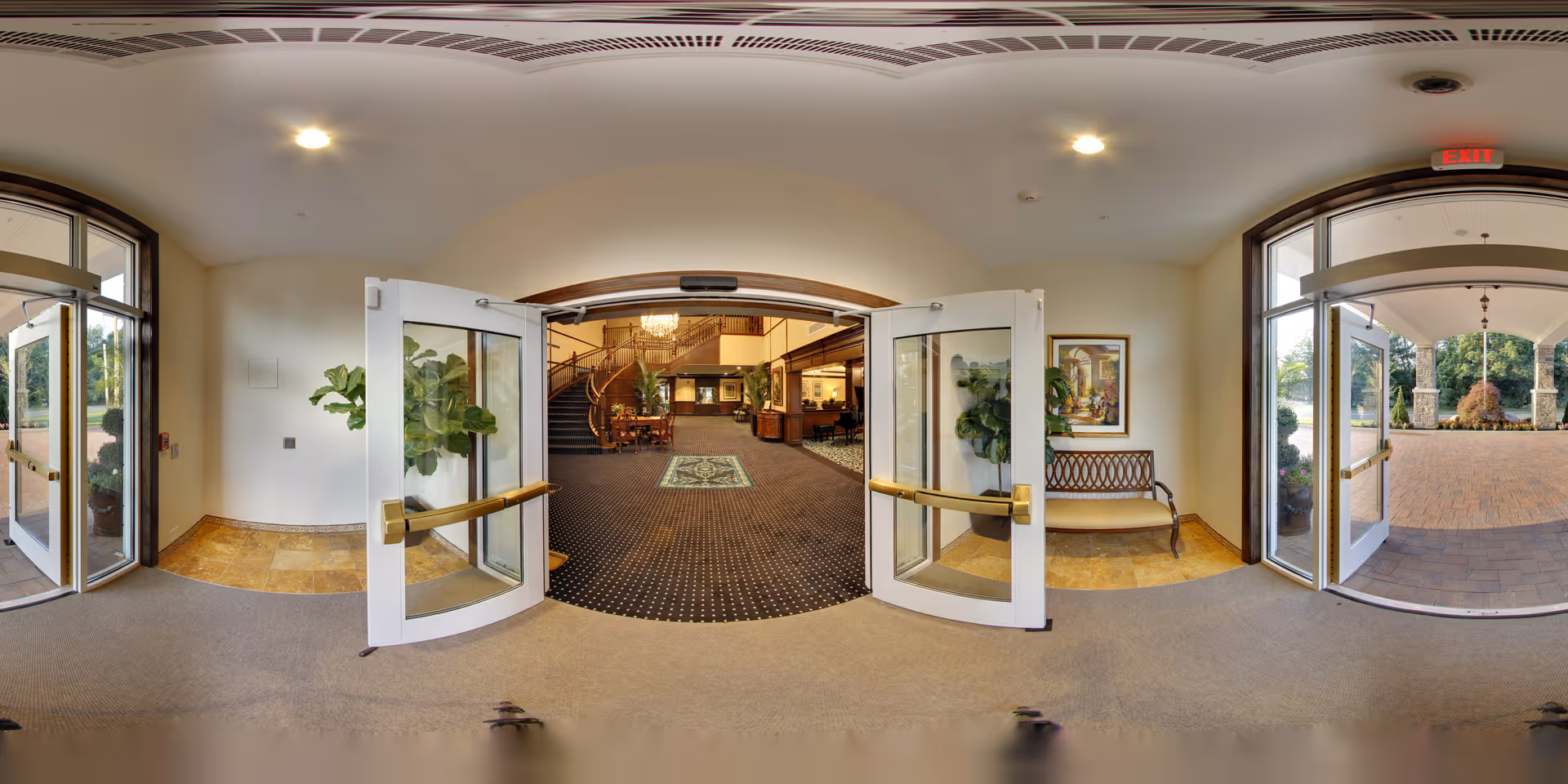 View from the entrance of Brandywine Mahwah by Monarch showing double glass doors opening into a spacious lobby area with carpeted floors, a staircase, plants, and seating. Outside, there is a covered driveway with brick paving and stone pillars.