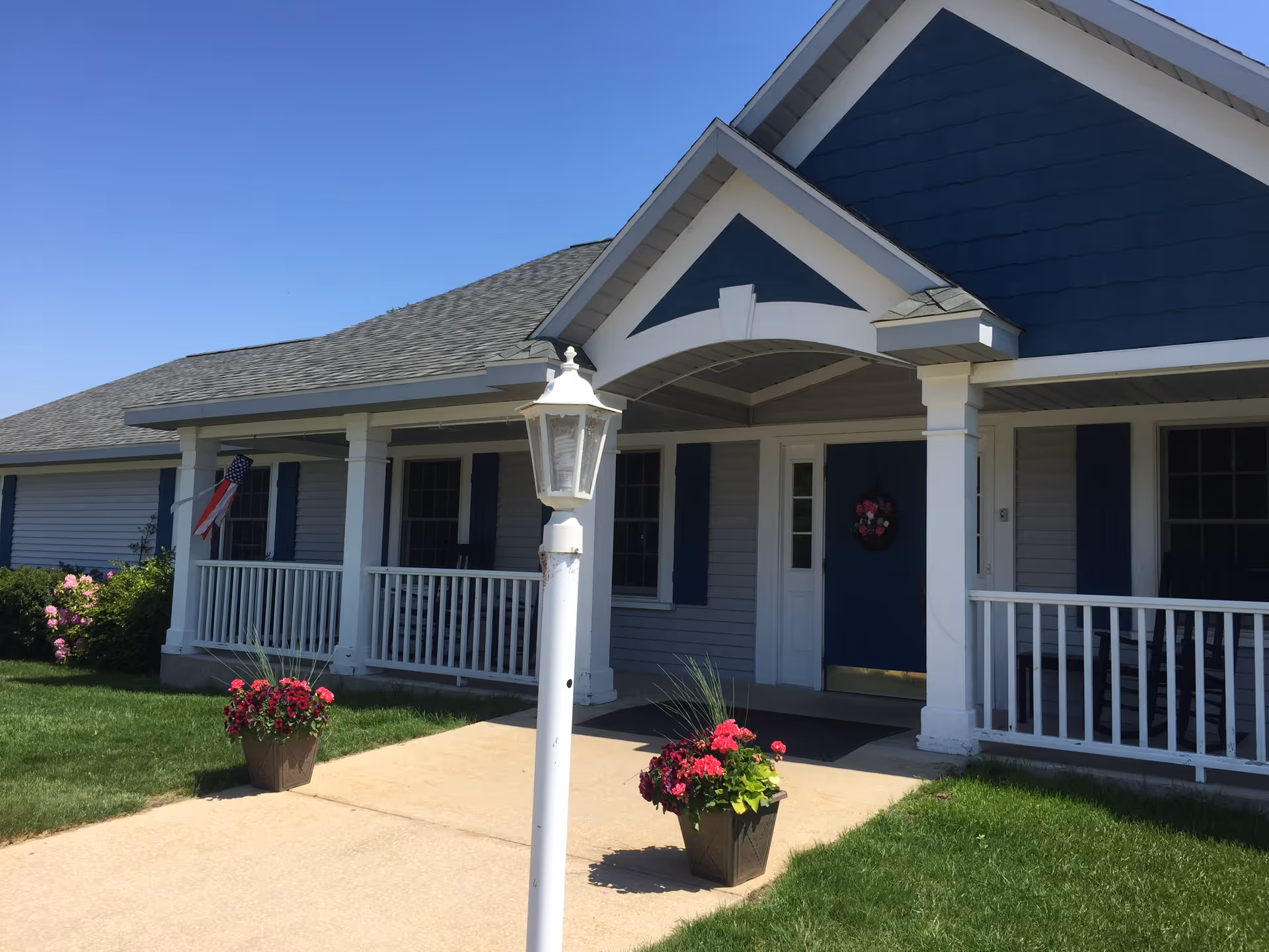 Front exterior view of a single-story building with a covered porch, white railings, and blue shutters. There are two flower pots with pink flowers on either side of the walkway leading to the entrance. An American flag is displayed near one window, and a white lamp post is positioned near the walkway.