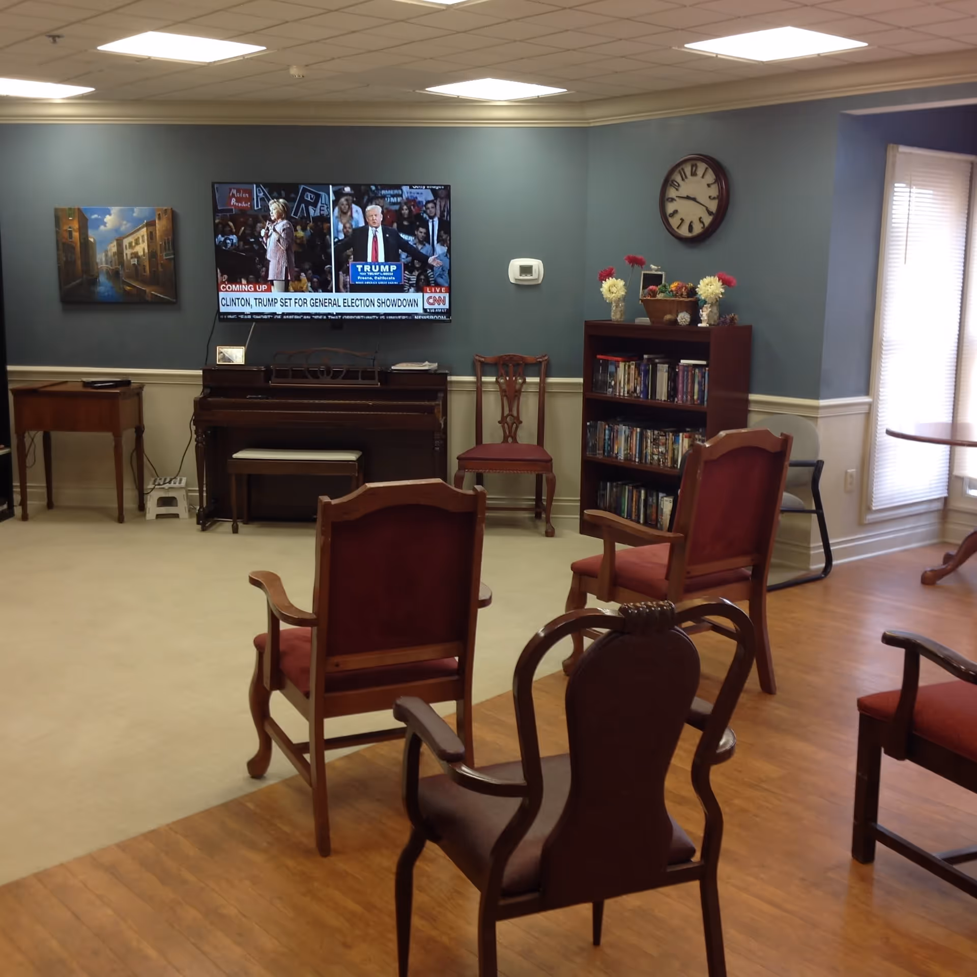 A common area in a senior living facility with several wooden chairs arranged facing a wall-mounted television showing a news broadcast. The room has a piano, a bookshelf filled with books and DVDs, a clock on the wall, and a window with blinds letting in natural light. The walls are painted blue with white trim, and the floor is a combination of carpet and wood.