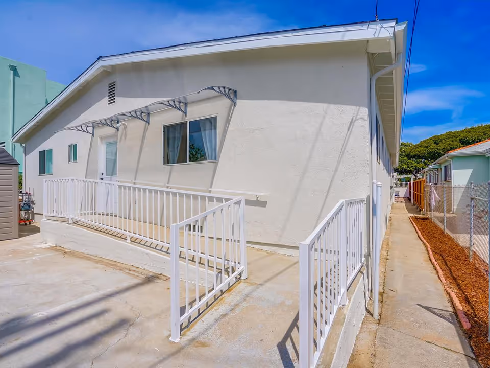 Exterior view of a single-story building with white walls and a white metal railing along a concrete ramp leading to a door. There is a window with curtains next to the door and a clear blue sky above. A narrow concrete pathway runs alongside the building, bordered by a chain-link fence and mulch on one side.