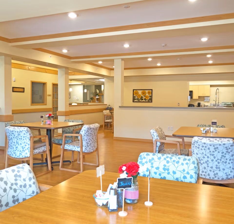 Bright communal dining area with wooden tables, floral-upholstered chairs, small table decorations, and a kitchen pass-through.