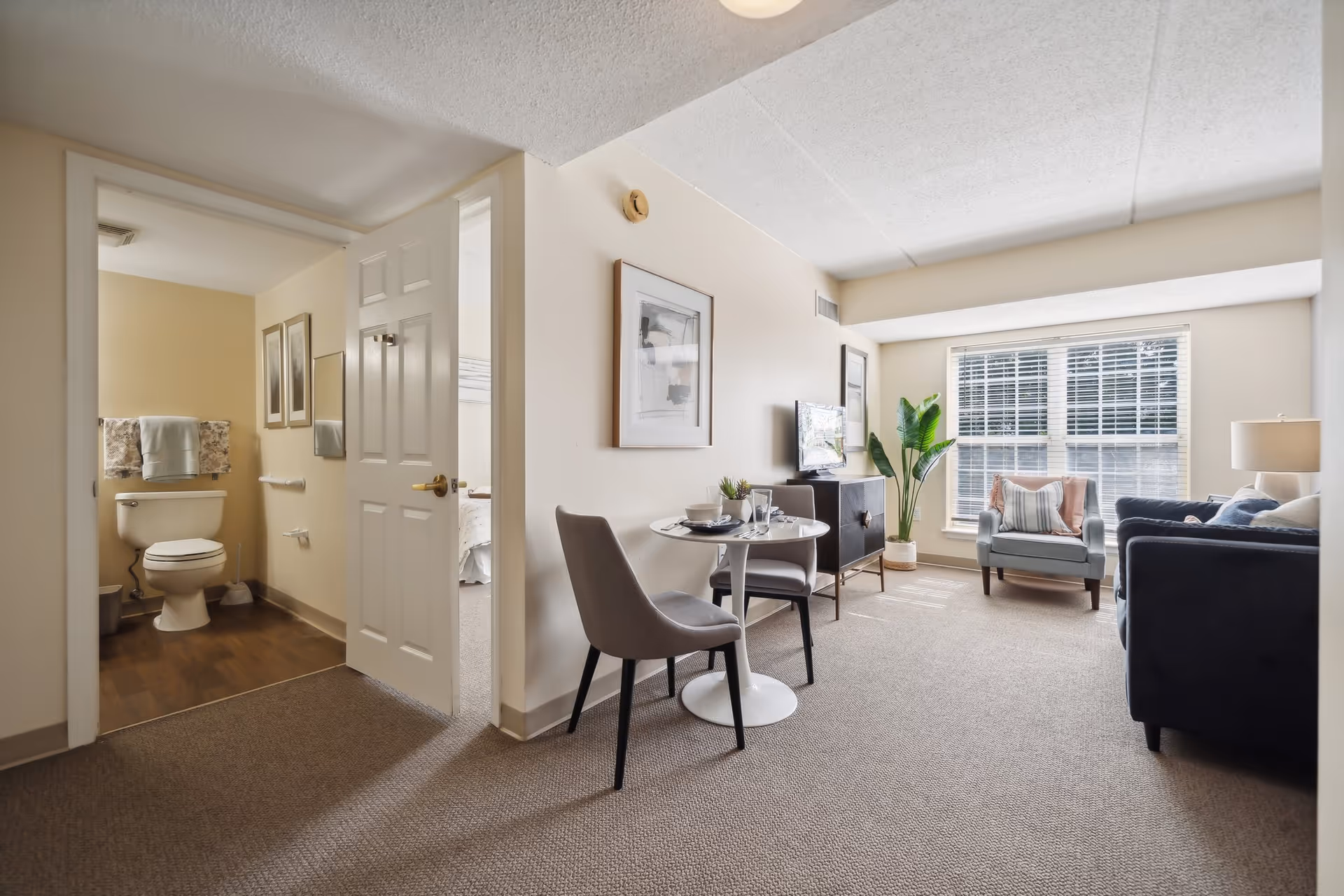 Cozy living room and small dining nook with chairs, TV, large window, and an open doorway showing a bathroom.