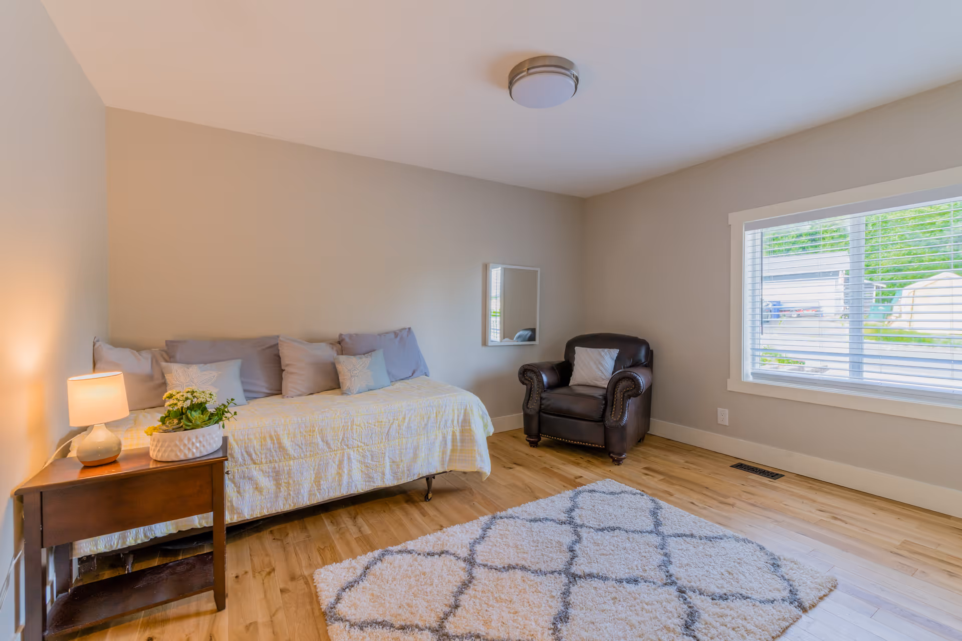 A cozy bedroom with a daybed covered in a light yellow quilt and several pillows, a wooden side table with a lamp and a potted plant, a dark brown leather armchair with a pillow, a large window with white blinds, a light fixture on the ceiling, and a patterned area rug on the wooden floor.
