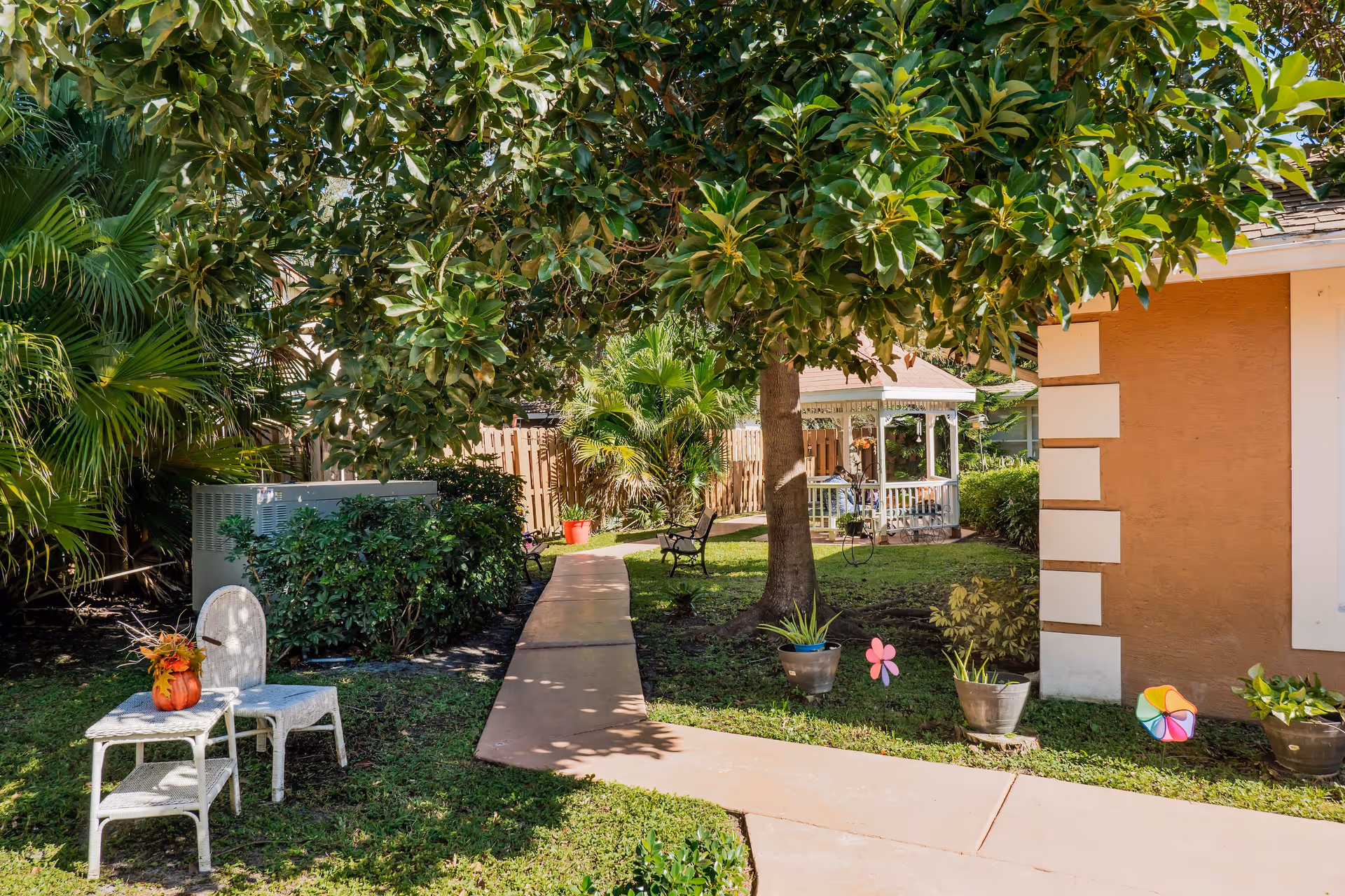 A sunny outdoor garden area with a paved walkway leading to a white gazebo. The scene includes green trees and bushes, a brown building wall on the right, two white wicker chairs with a small table holding a pumpkin decoration on the left, and several potted plants and colorful pinwheels along the path.
