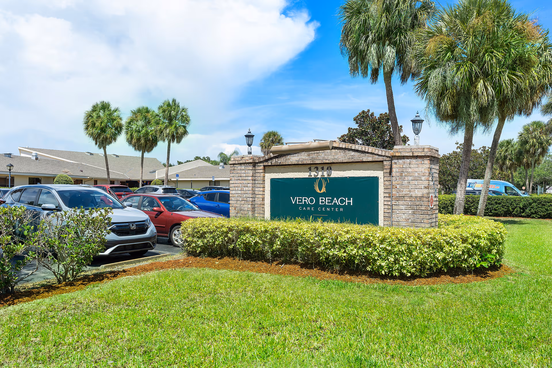 Stone entrance sign reading 'Vero Beach Care Center' on a landscaped lawn with parked cars and palm trees in the background.