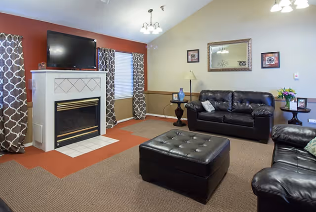 A cozy living room with two black leather sofas, a matching black leather ottoman, a fireplace with a TV mounted above it, patterned curtains on the windows, a floor lamp, side tables with decorative items, and framed artwork on the walls.