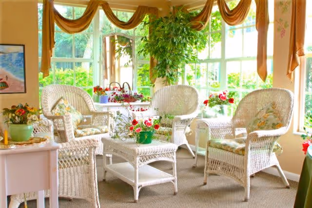 A bright and cozy sunroom with white wicker furniture including chairs and a table, decorated with floral cushions and various potted plants and flowers. Large windows with golden drapes allow natural light to fill the room.