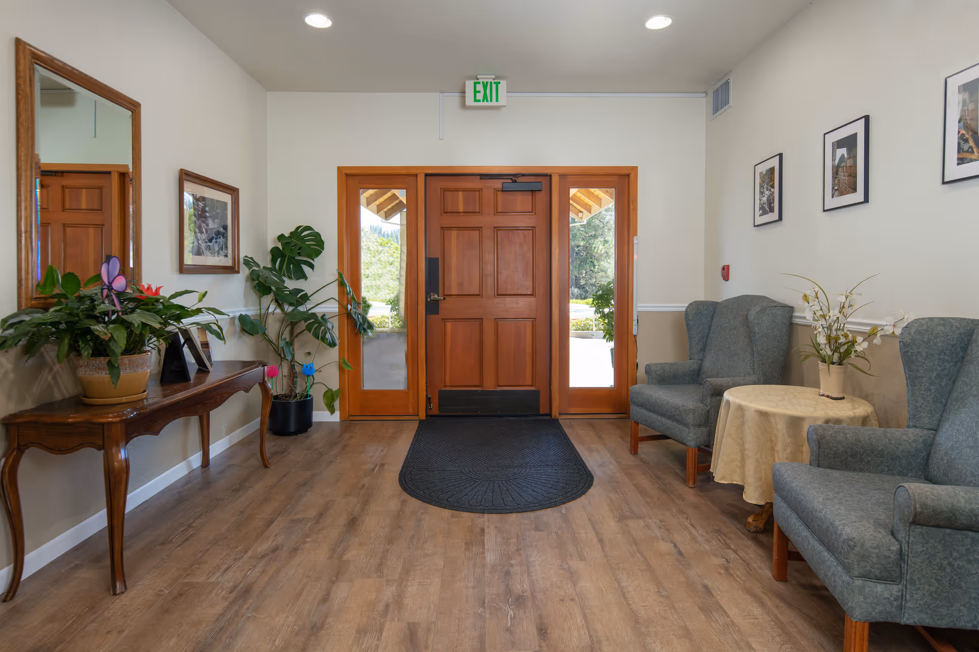 A cozy entrance area with a wooden door flanked by glass panels. On the left side, there is a wooden console table with potted plants and a mirror above it. On the right side, there are two upholstered armchairs with a small round table covered with a beige cloth and a flower arrangement. The floor is wooden, and there is a black mat in front of the door. The walls are decorated with framed pictures.
