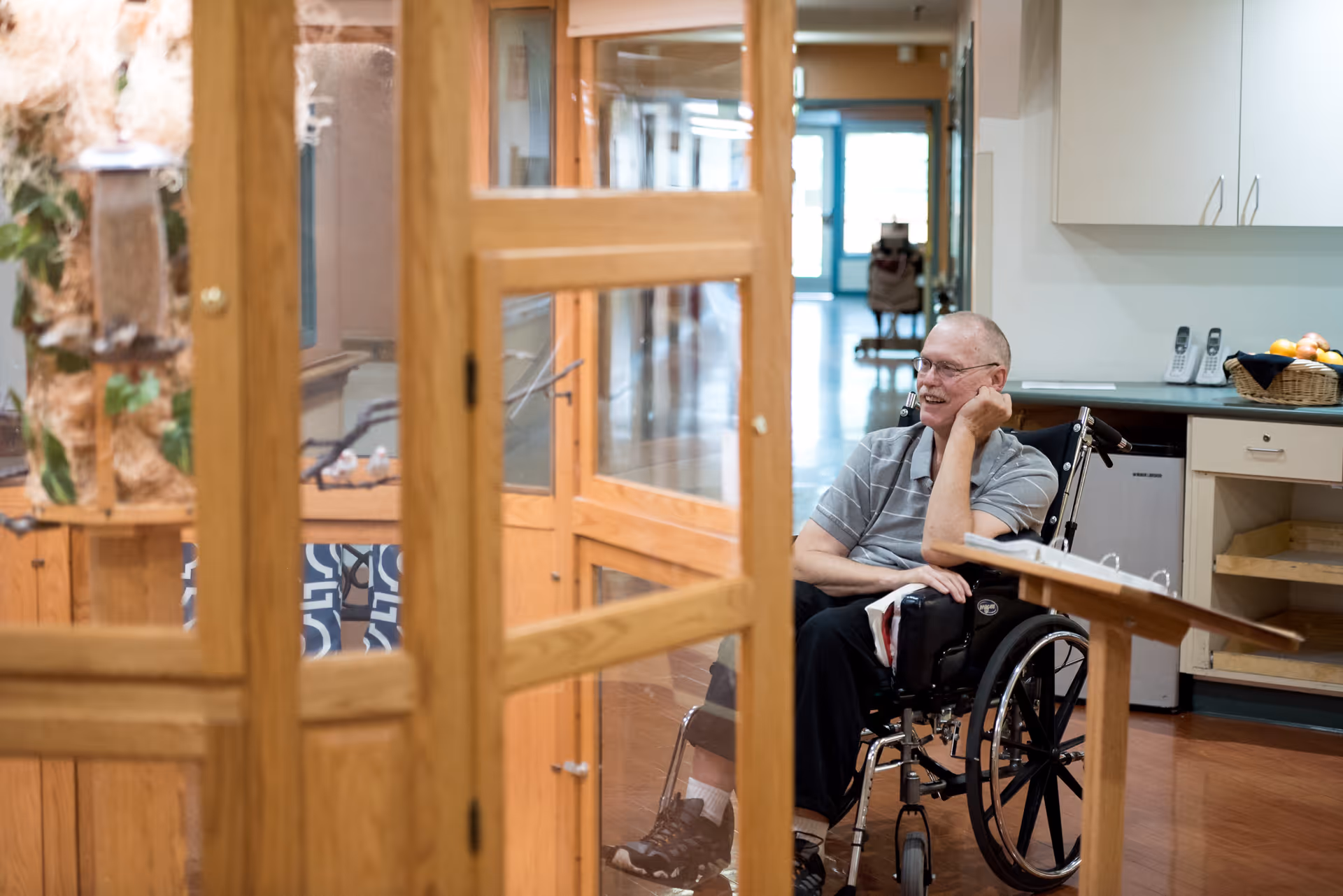 An elderly man in a wheelchair is sitting and smiling inside a room with wooden framed glass doors. The room has a kitchen area with white cabinets, a countertop with a basket of fruit, and cordless phones. The floor is wooden, and the background shows a hallway with natural light coming through a door.