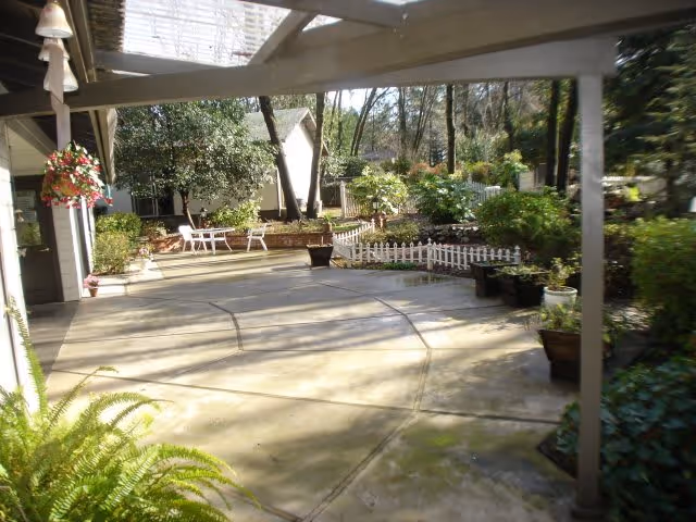 A spacious outdoor patio area with concrete flooring, surrounded by various plants and greenery. There are white chairs and a small table in the background near a garden bed with a white picket fence. The patio is partially covered by a pergola structure, and hanging flower baskets and potted plants add to the garden ambiance.