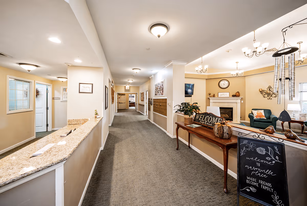 Interior view of a senior living facility hallway with a reception desk on the left and a cozy sitting area on the right featuring a fireplace, armchairs, and decorative items including a welcome sign and a chalkboard sign that reads 'welcome to masonville place where friends become family.' The hallway is carpeted and well-lit with ceiling lights.