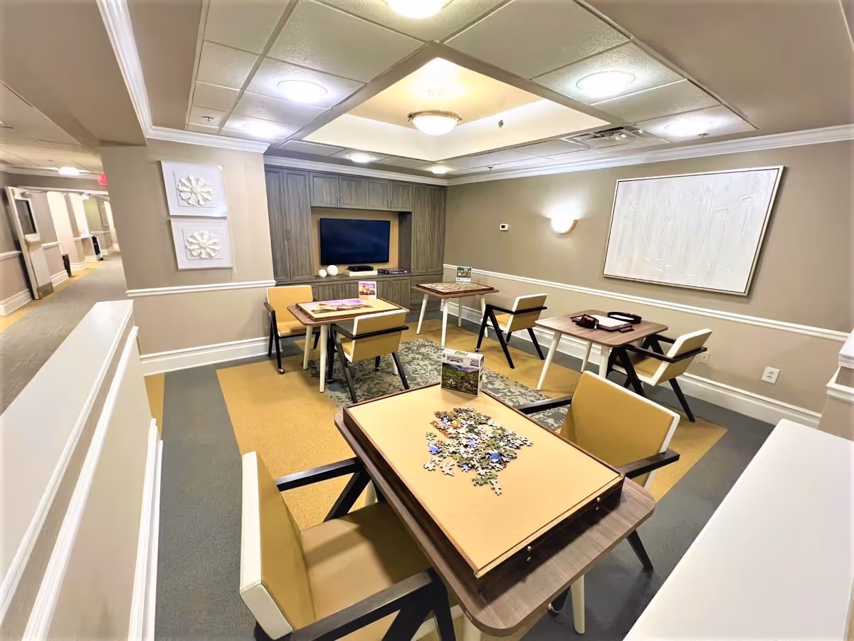 A well-lit common area in an assisted living facility with four small tables, each surrounded by chairs. One table has a partially completed jigsaw puzzle on it. The room features neutral-colored walls, a built-in wooden entertainment center with a flat-screen TV, decorative wall art, and a patterned rug under two of the tables. A hallway is visible to the left.