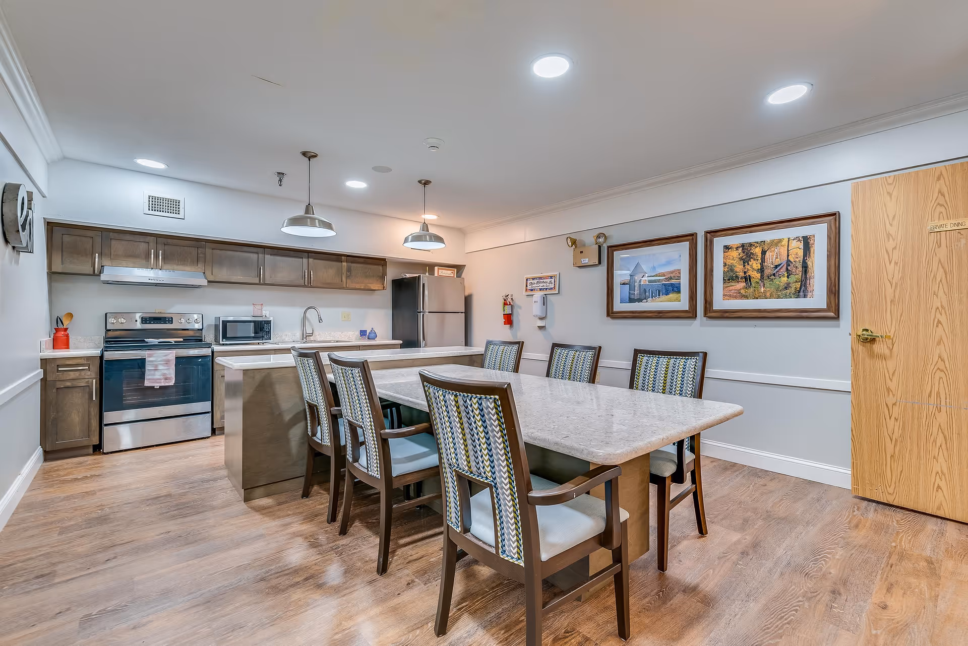 Communal dining area with a long table and upholstered chairs next to a modern kitchen island and stainless steel appliances.