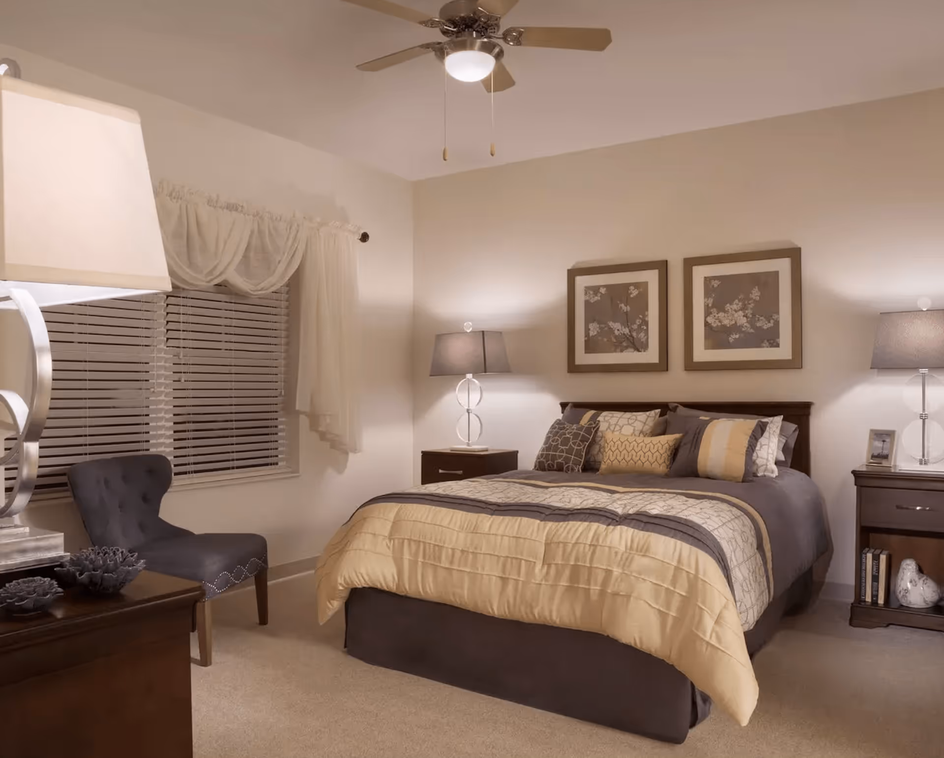 A cozy bedroom with a large bed covered in a beige and gray comforter and multiple decorative pillows. There are two matching nightstands on either side of the bed, each with a lamp. Above the bed are two framed floral artworks. A gray upholstered chair is positioned near a window with white blinds and sheer curtains. The room is softly lit by the lamps and a ceiling fan with a light.