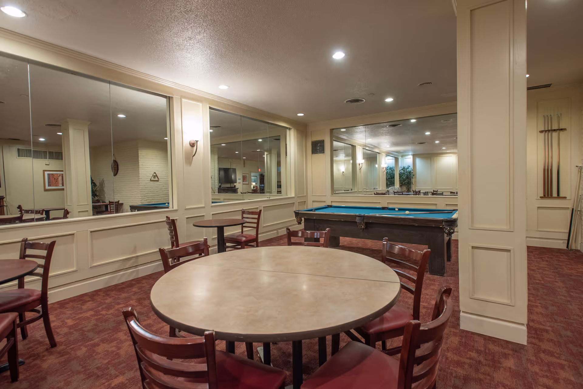 Interior room with round tables and chairs arranged around them, a pool table with billiard balls, and large mirrors on the walls reflecting the room. The room has beige walls, red carpet, and soft lighting from wall sconces and ceiling lights.
