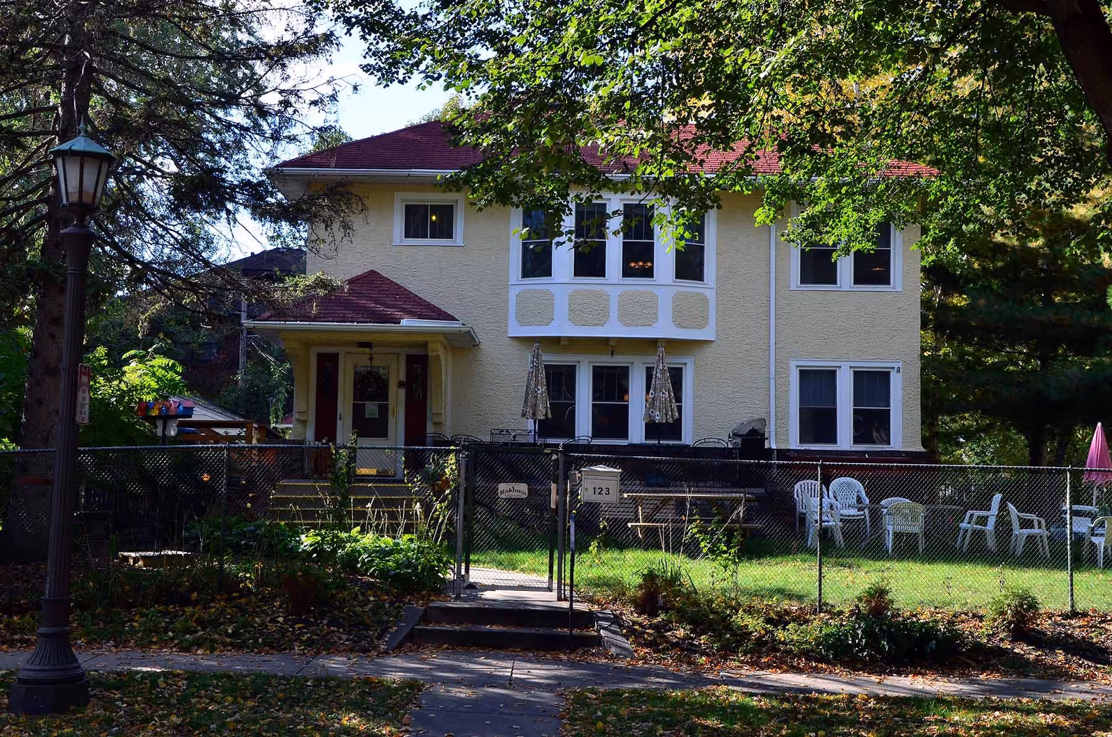 Front exterior view of a two-story yellow house with a red roof, surrounded by trees and a fenced yard with white plastic chairs and umbrellas. A street lamp is visible on the left side of the image.