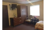 Small furnished bedroom with a dresser and wardrobe, a wall-mounted TV, two chairs by a window, and a partially visible bed.