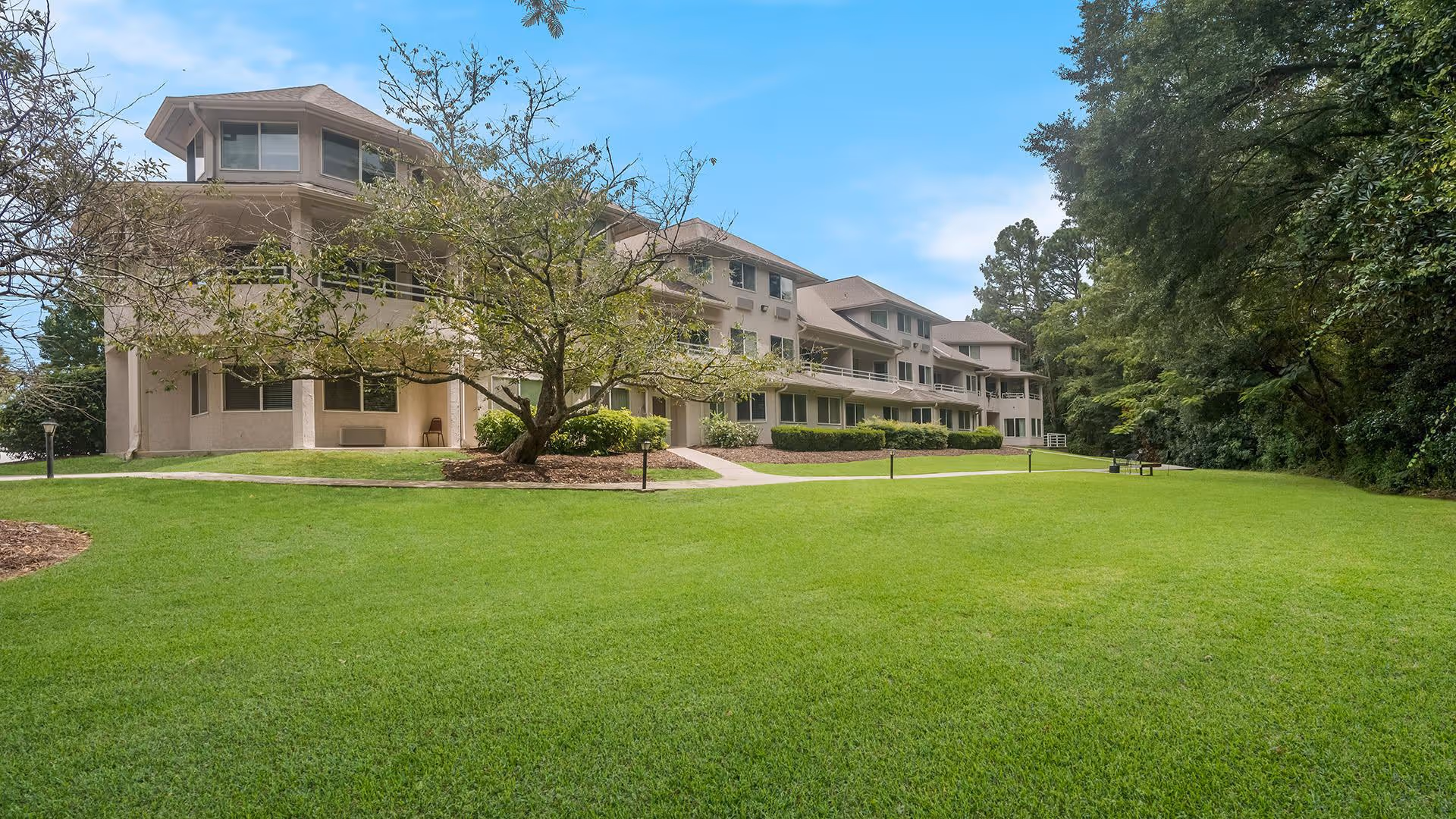 Exterior view of a multi-story senior living facility building with beige walls and multiple balconies, surrounded by a well-maintained green lawn and trees under a blue sky.