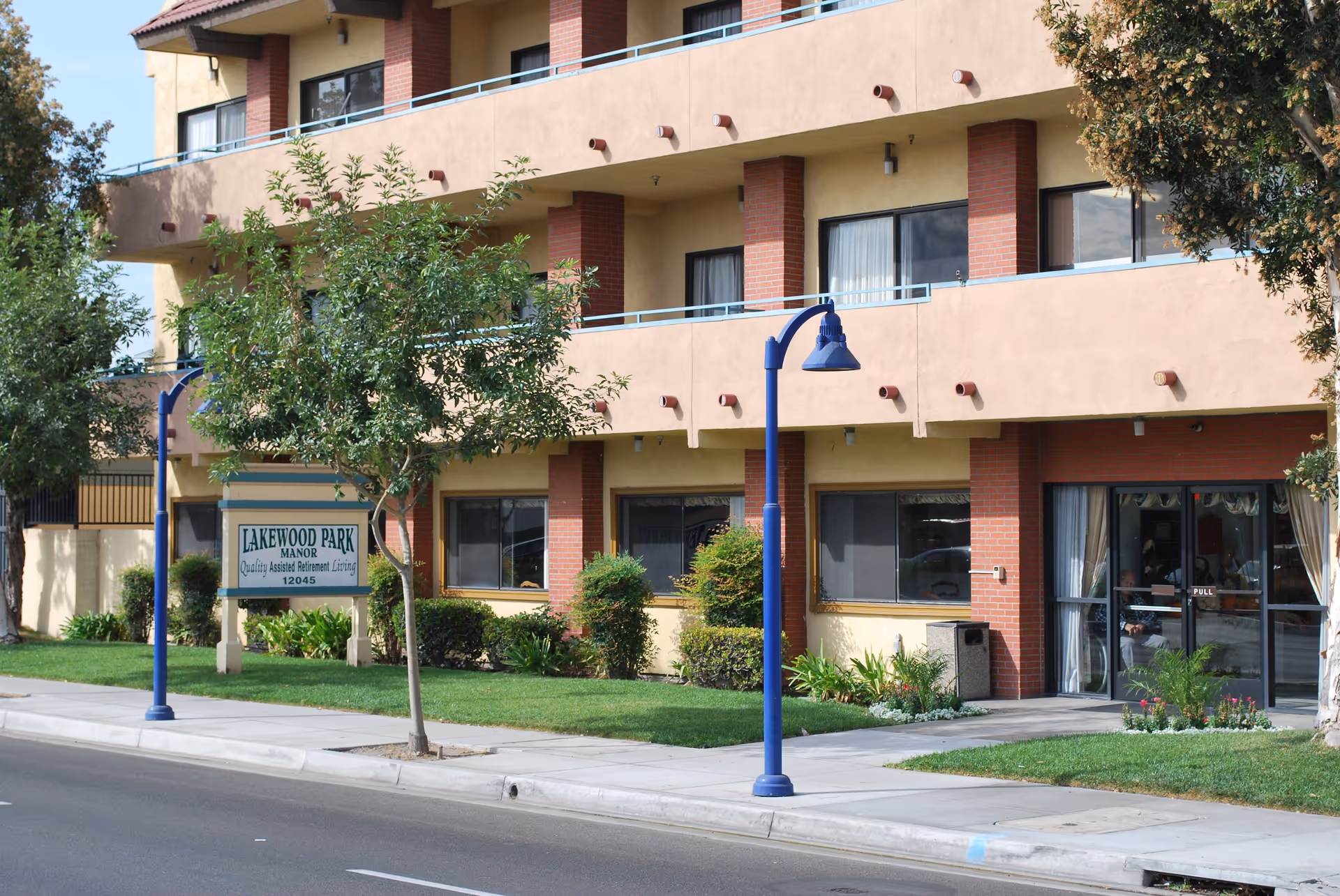 Exterior view of Lakewood Park Manor, a multi-story assisted retirement living facility with beige walls and red brick accents. The building has balconies with blue railings, large windows, and a glass entrance door. There are green trees, shrubs, and a well-maintained lawn in front of the building along a sidewalk and street. A sign near the sidewalk reads 'Lakewood Park Manor Quality Assisted Retirement Living 12045'.