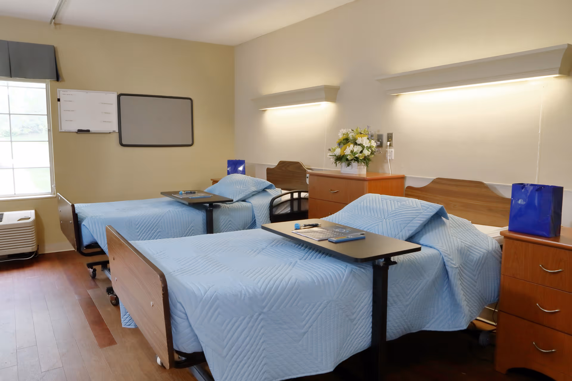 Two twin hospital-style beds with blue bedspreads in a bright shared senior room with bedside cabinets, over-bed tables, and a window.