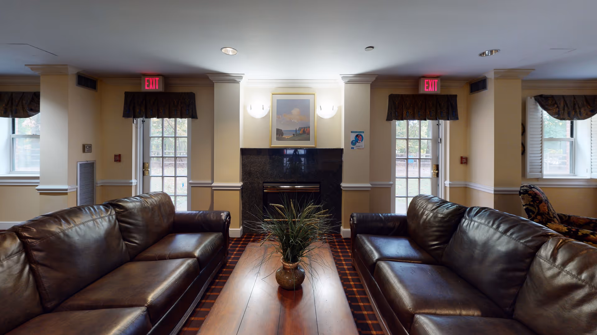 Interior view of a cozy living room in a senior living facility featuring two dark brown leather sofas facing each other with a wooden coffee table in between. A potted plant sits on the coffee table. The room has a fireplace with a framed painting above it, flanked by two wall sconces. There are two glass doors with exit signs above them and windows with valances on either side, allowing natural light to enter.