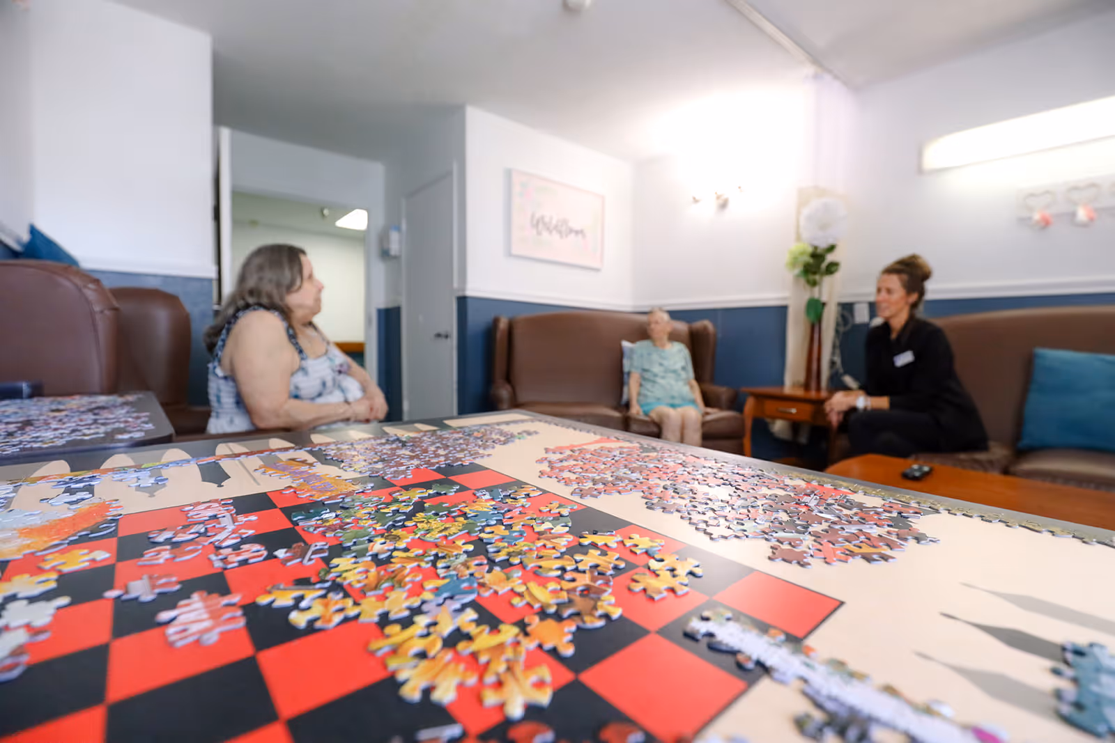 A group of three people sitting and talking in a living room area of a senior living facility. In the foreground, there is a table with an incomplete jigsaw puzzle. The room has brown leather chairs, a wooden side table with a flower vase, and light blue walls with white trim.