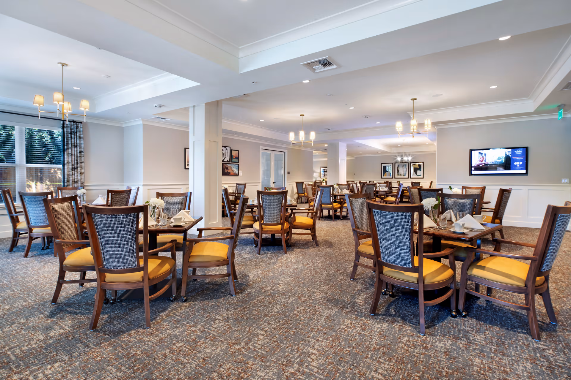 Spacious dining room with multiple round tables and wooden chairs with yellow cushions, chandeliers, and a wall-mounted TV.