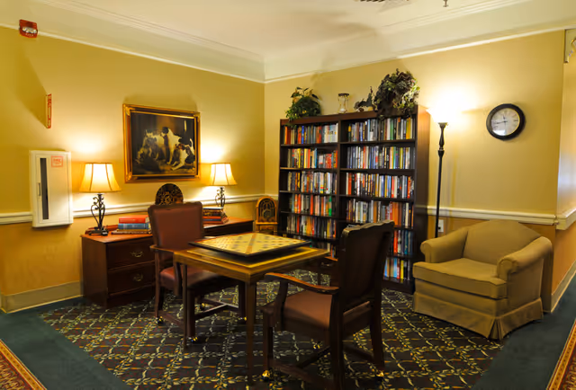 A cozy interior room with a small table featuring a chessboard, surrounded by two chairs. There is a bookshelf filled with books against the wall, a comfortable armchair, two table lamps on a wooden sideboard, a floor lamp, a wall clock, and a framed painting of dogs on the wall.