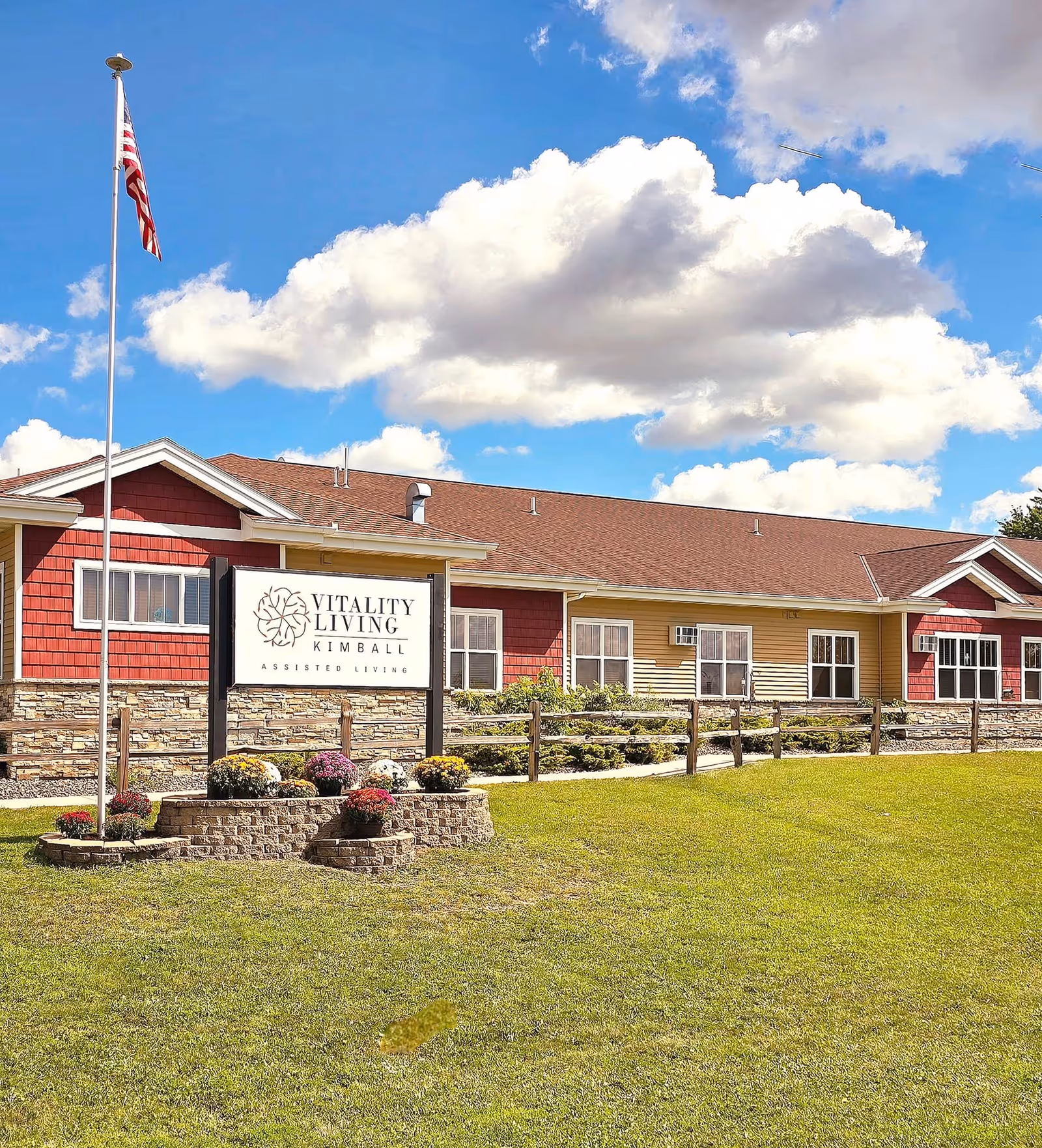 Exterior view of Vitality Living of Kimball assisted living facility with a manicured lawn, a flagpole with an American flag, a sign displaying the facility's name, and a building with red and yellow siding under a partly cloudy blue sky.