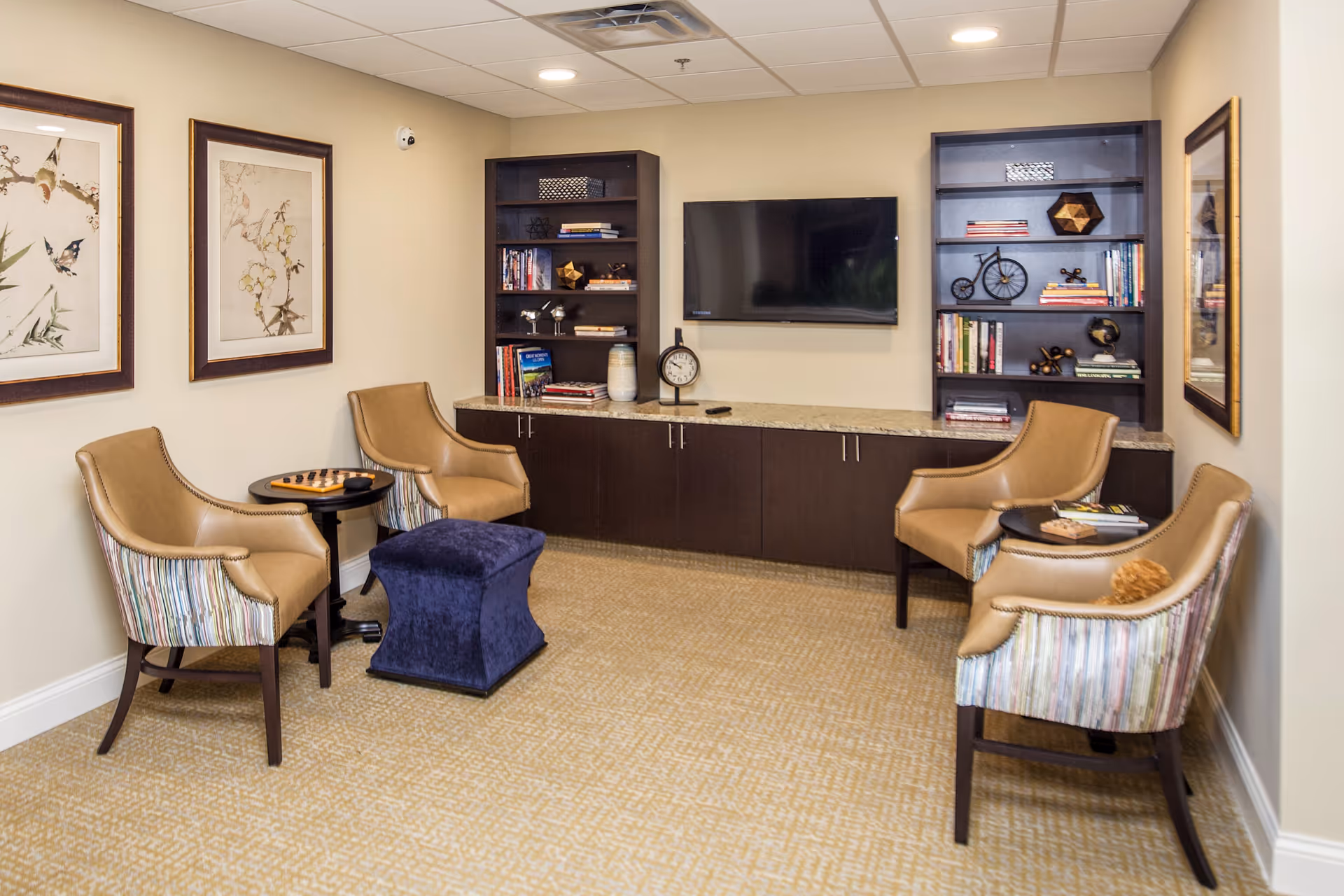 A cozy senior living common area with four tan and striped armchairs arranged around two small round tables and a blue ottoman. The room features beige walls with framed artwork, a wall-mounted flat-screen TV, and dark wood built-in shelves and cabinets filled with books and decorative items.