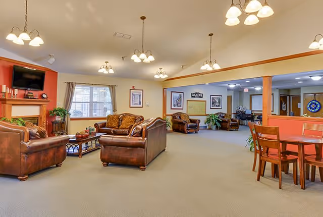 A spacious living room area in a senior living facility with leather sofas and armchairs arranged around a coffee table. The room features a fireplace with a TV mounted above it, several hanging light fixtures, large windows with curtains, and framed artwork on the walls. There is also a wooden dining table with chairs on the right side and plants placed around the room.