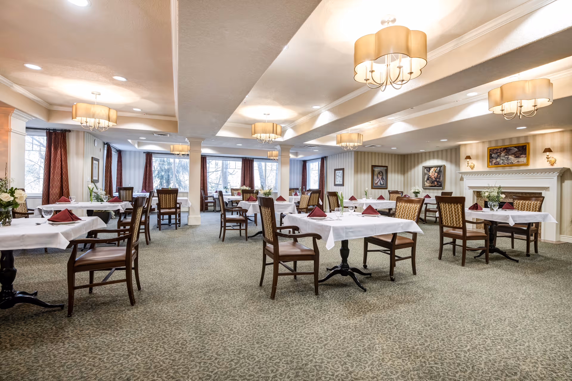 A spacious dining room with multiple tables covered in white tablecloths, each set with folded red napkins, glassware, and floral centerpieces. The room features patterned carpet, large windows with red curtains, framed artwork on the walls, and elegant ceiling light fixtures. There is a white fireplace on the right side of the room.