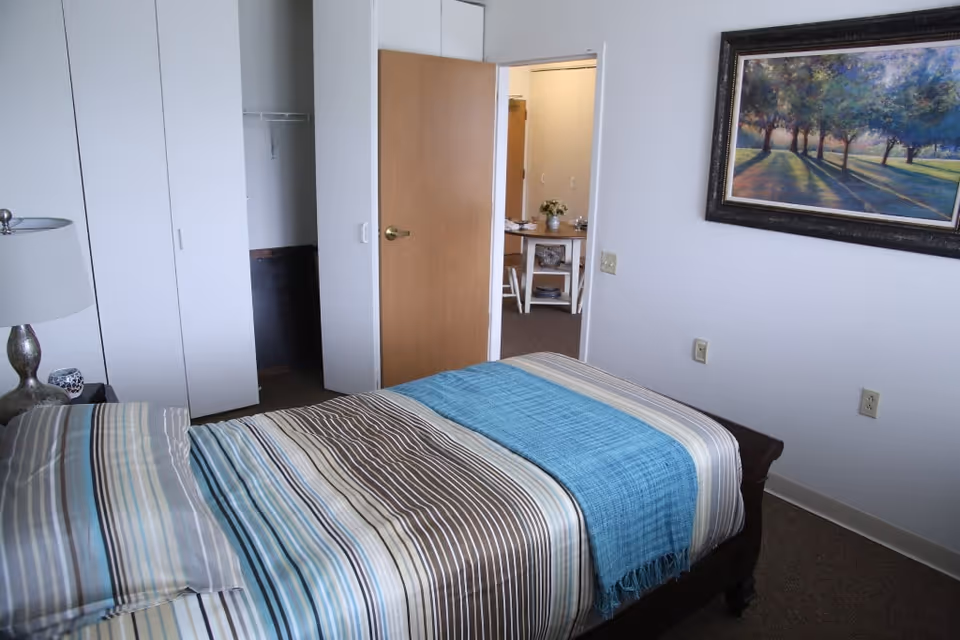 A bedroom with a bed covered in striped bedding and a blue throw blanket. There is a bedside table with a lamp and a small decorative item. The room has white walls, a framed painting of trees on one wall, a closet with white doors, and an open wooden door leading to another room with a table and chairs.