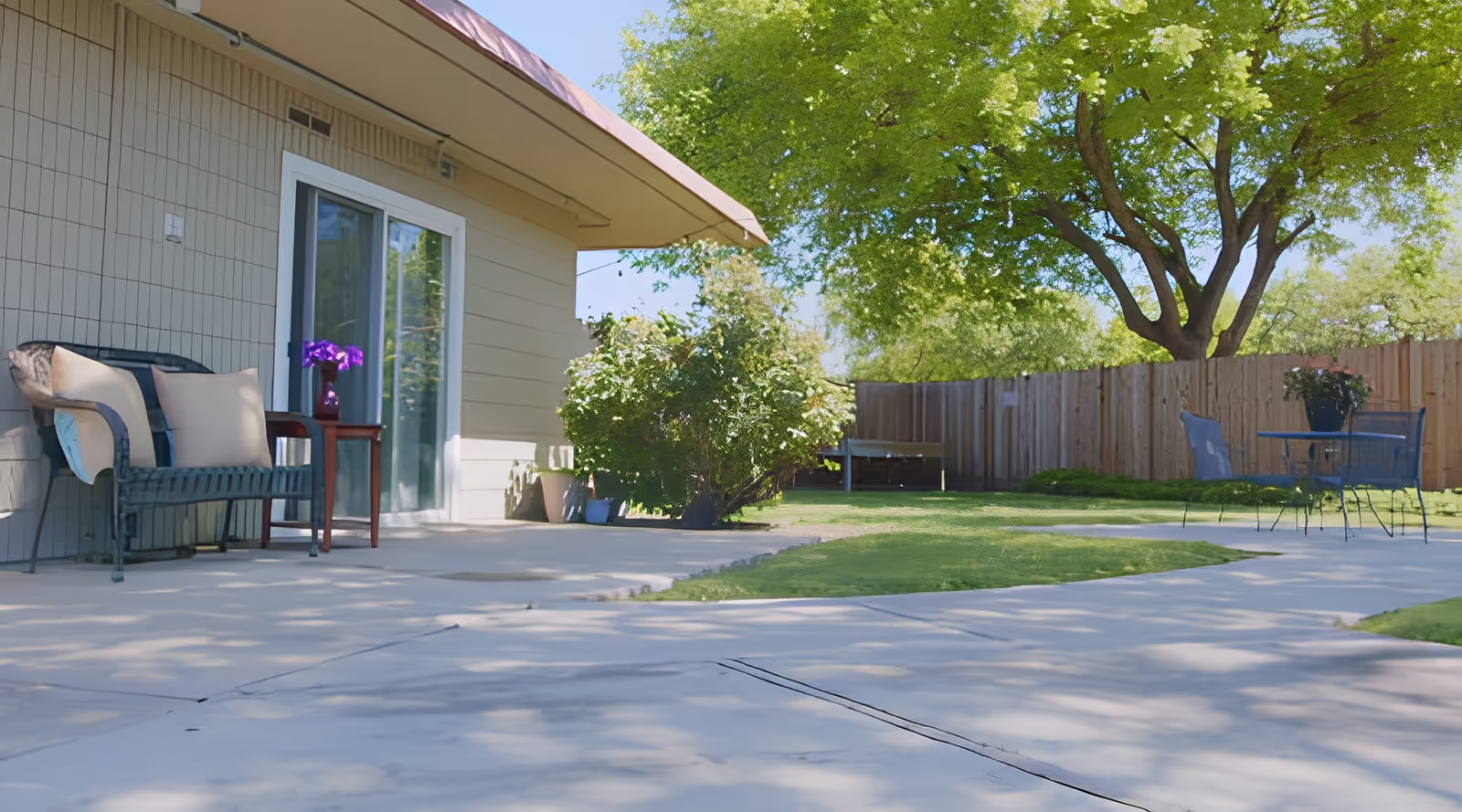Outdoor patio area with a bench and cushions next to a building with sliding glass doors. There is a small table with a vase of purple flowers beside the bench. The patio extends to a grassy yard with a wooden fence, a large tree providing shade, and a metal table with chairs and a potted plant.