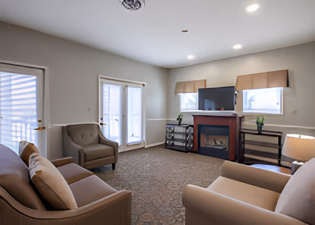 A cozy living room with beige walls and carpeted floor. The room features a brown sofa with cushions, two beige armchairs, a fireplace with a TV mounted above it, two windows with beige valances, and two small black side tables with plants. There is a door and a set of French doors letting in natural light.