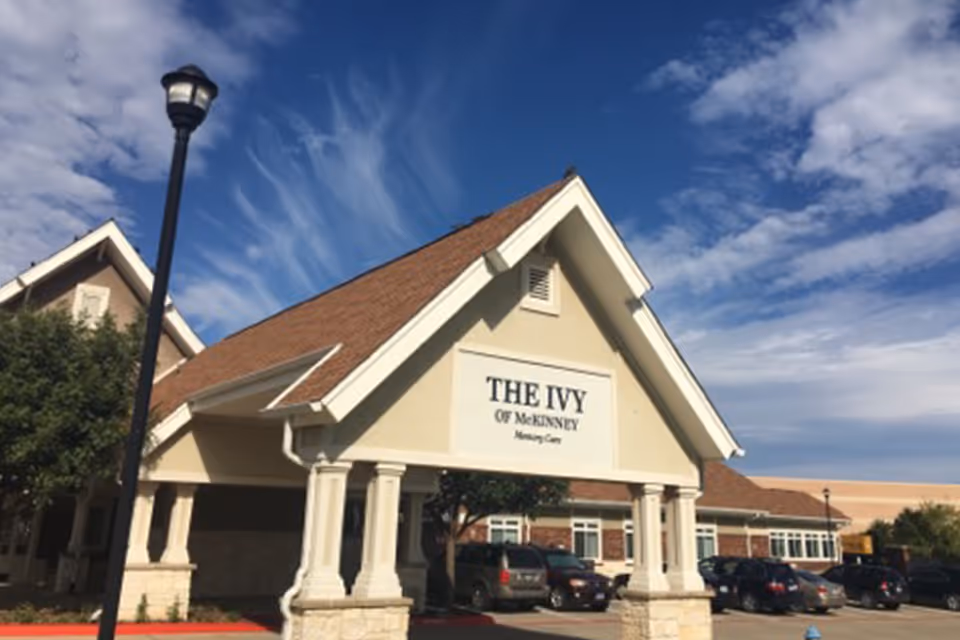 Front entrance canopy of The Ivy of McKinney senior living building with parked cars under a blue sky.
