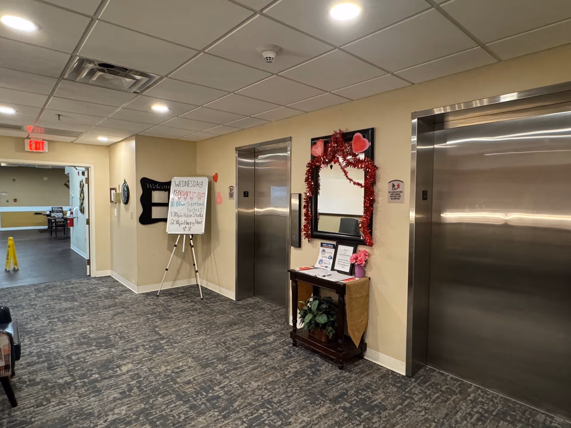 Interior hallway of a senior living facility with two stainless steel elevators on the right side. A small wooden table with a mirror above it decorated with red garland and heart-shaped decorations is placed between the elevators. A whiteboard on an easel stands near the elevators displaying the day's schedule including Super Bowl posters, Bible study, and happy hour. The hallway has carpeted flooring and beige walls, with a doorway leading to another room visible on the left.