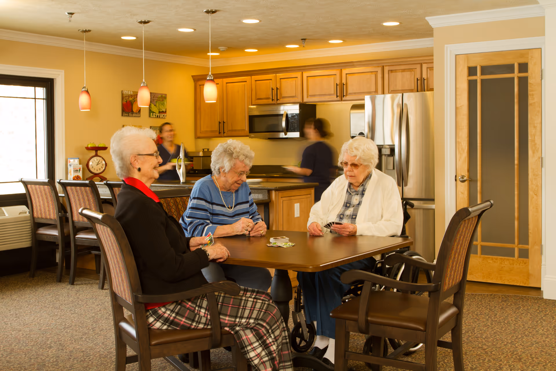 Three elderly women sitting around a wooden table playing cards in a well-lit kitchen area with wooden cabinets, stainless steel appliances, and two staff members blurred in the background.