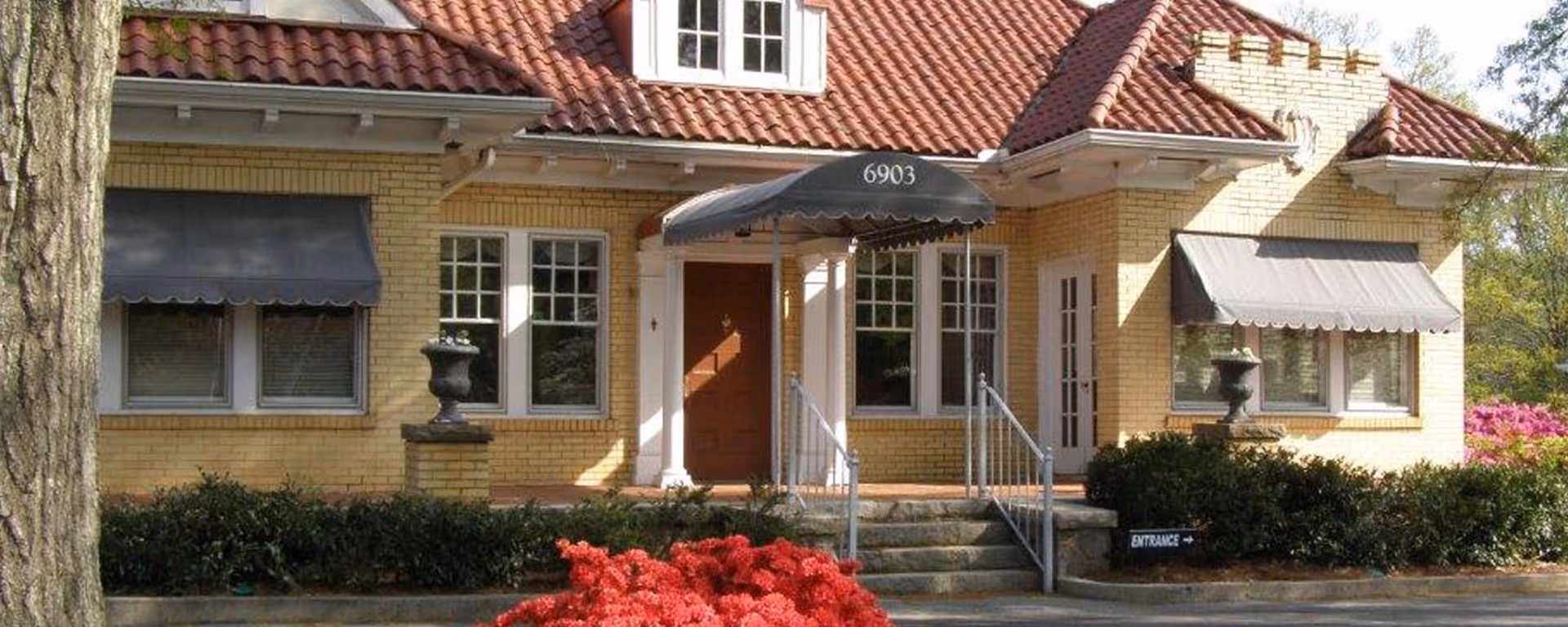 Exterior view of a yellow brick building with a red tiled roof, featuring a central entrance with a black awning displaying the number 6903. There are windows with gray awnings on either side of the entrance, bushes and red flowers in front, and a small sign indicating the entrance direction.
