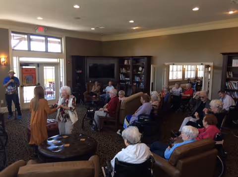 Seniors and staff gathered in a community lounge watching a performer while two women dance near the center.