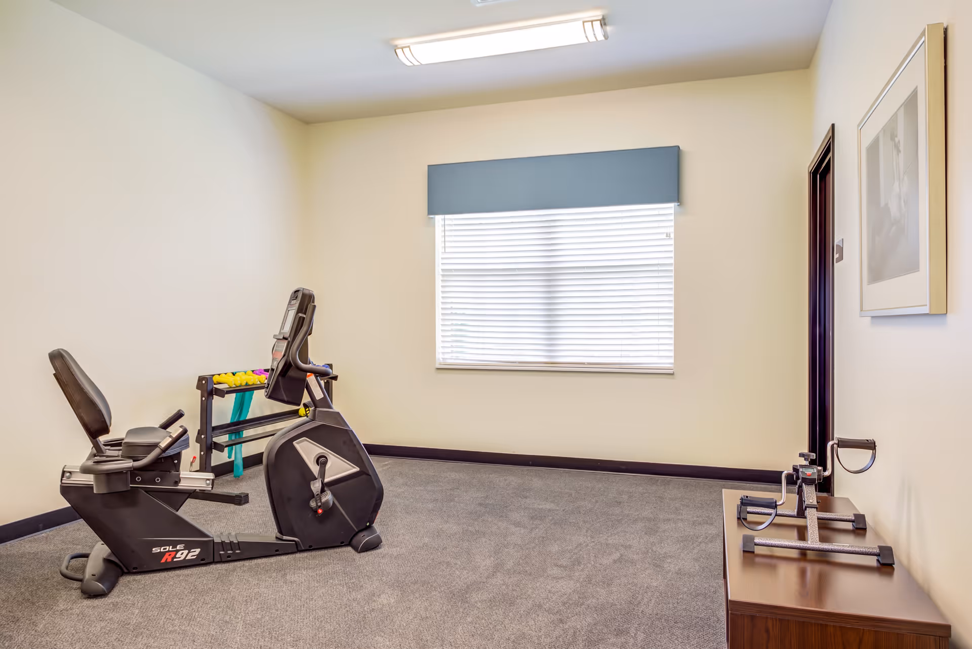Small exercise room with a recumbent stationary bike, a rack of dumbbells and resistance bands, and a window with blinds.