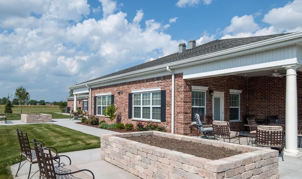 Outdoor patio area of a senior living facility with brick exterior walls, white columns, multiple windows with shutters, outdoor seating including metal chairs and tables, and a raised stone planter bed. The sky is partly cloudy and the surrounding area includes a grassy lawn and some small trees.