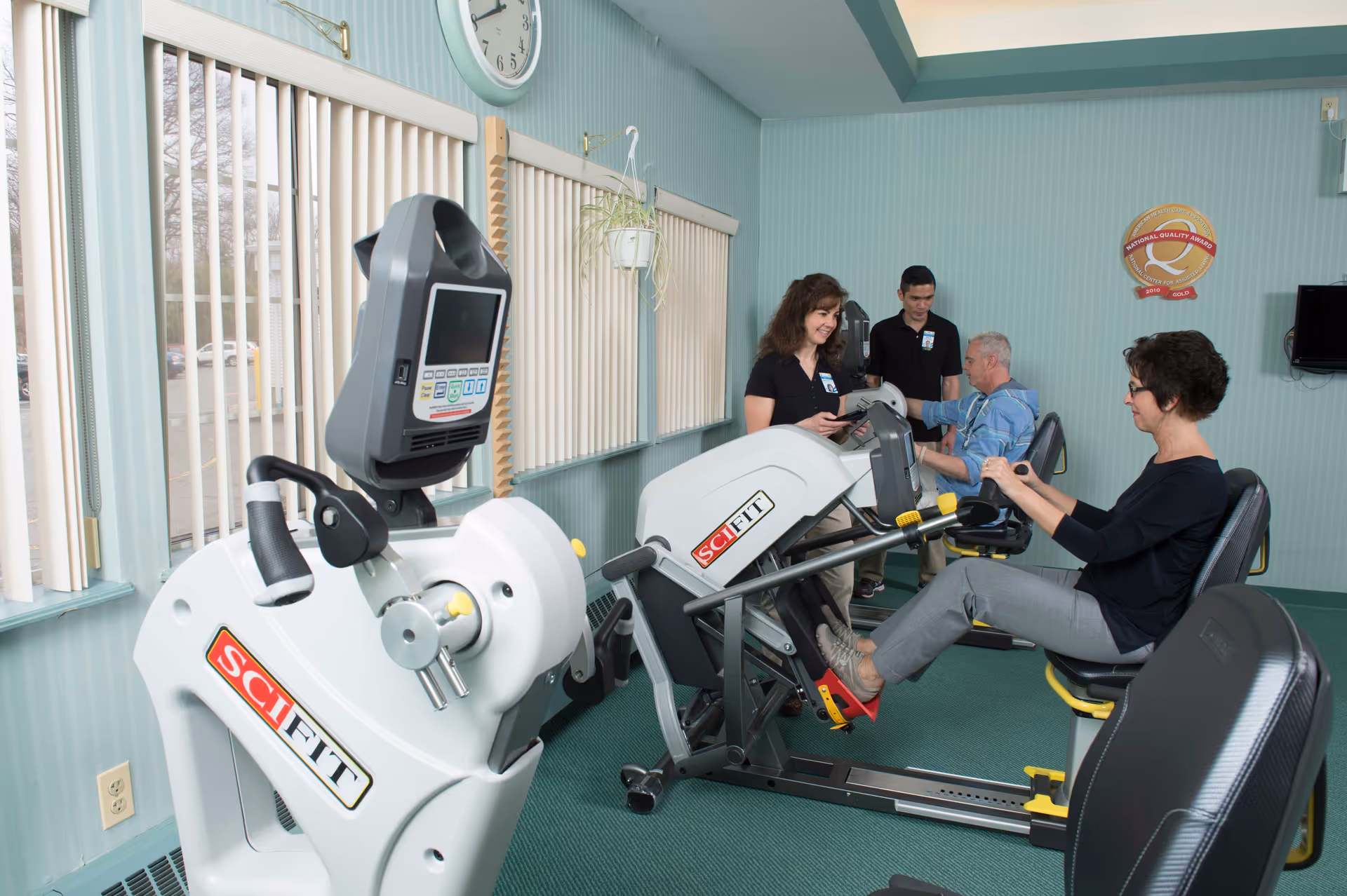 Seniors and staff using seated exercise machines in a mint-green activity/therapy room.