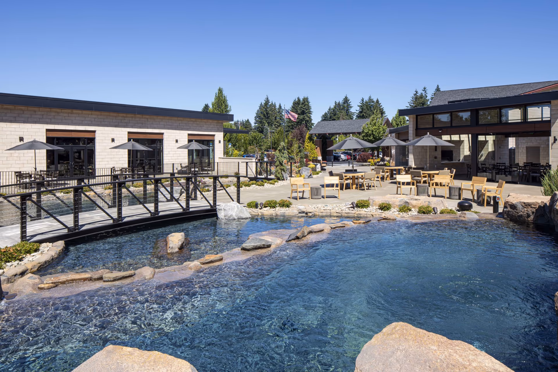 Outdoor patio area at Panorama facility featuring a clear blue pond with rocks, a small black metal bridge, multiple tables and chairs with umbrellas, surrounded by buildings and trees under a clear blue sky.