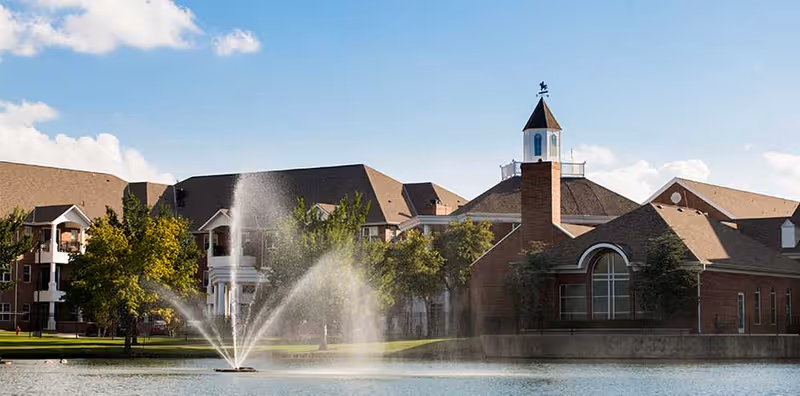 Brick senior living buildings with a cupola beside a pond and a fountain under a blue sky.