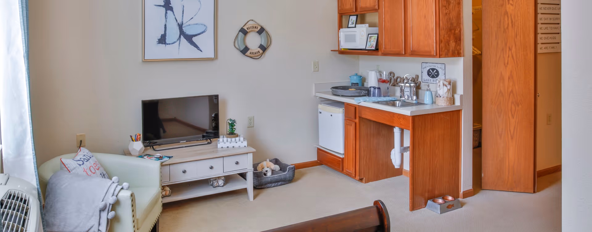 A cozy living area with a small white armchair draped with a gray blanket and a decorative pillow, a white TV stand holding a flat-screen television and small decor items, a kitchenette with wooden cabinets, a microwave, a mini fridge, a sink, and various kitchen items on the counter. There is a small dog bed with a stuffed dog toy on the floor near the kitchenette. A wooden door is partially open revealing a closet, and a wall decoration with family rules is visible in the hallway.