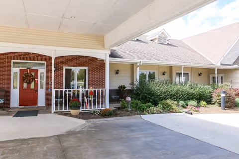 Covered entrance area of a senior living facility with a red front door decorated with a fall wreath, brick and beige siding exterior walls, potted plants, shrubs, and a concrete driveway.
